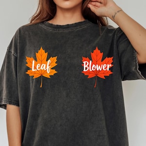Peut inclure: T-shirt gris foncé avec les mots "Leaf" et "Blower" encadrant deux feuilles d'érable orange et rouge. Le t-shirt a un aspect vintage et délavé, avec une coupe ample, parfait pour l'automne.
