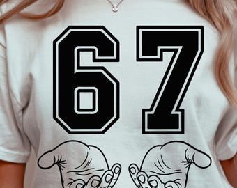 May include: White t-shirt featuring the number 67 in black, outlined in white. Below the numbers are two black and white illustrations of open hands. The shirt is worn by a person.