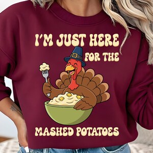 Peut inclure: Sweat-shirt bordeaux avec l'inscription "I'M JUST HERE FOR THE MASHED POTATOES" et une illustration d'une dinde de dessin animé portant un chapeau de pèlerin, tenant une fourchette avec de la purée de pommes de terre dans un bol vert.
