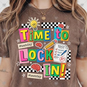 May include: Brown t-shirt with a colorful graphic that says "Time to Lock In!" The design includes a lightbulb, brain, pencil, test paper, and stars. The words are in various bright colors, with the hashtag #testday.