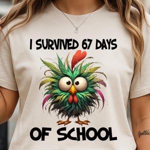 Peut inclure: T-shirt beige avec l'inscription "I SURVIVED 67 DAYS OF SCHOOL". Le t-shirt présente un dessin de poulet de dessin animé avec des détails verts, rouges et jaunes. Le poulet a de grands yeux et est centré sur le t-shirt.