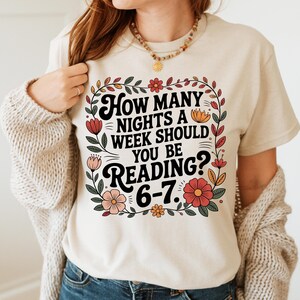 May include: Beige t-shirt with the text "How Many Nights A Week Should You Be Reading? 6-7." The text is surrounded by a floral design with red, pink, and yellow flowers and green leaves. The shirt is a casual style.