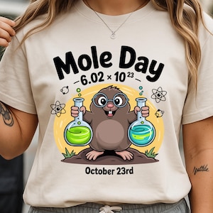 May include: Beige t-shirt with a graphic celebrating Mole Day. The design features a cartoon mole holding flasks with green and blue liquids, with the text "Mole Day - 6.02 x 10^23" and "October 23rd".
