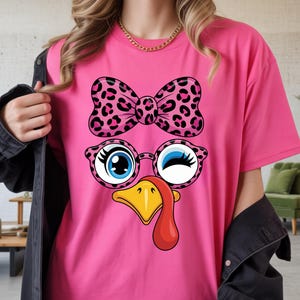 Peut inclure: T-shirt rose vif avec un motif de visage de dinde de dessin animé. La dinde porte un nœud papillon et des lunettes à imprimé léopard, des yeux bleus, un bec jaune et un caroncule rouge. Design coloré.