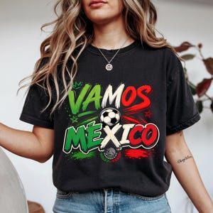 May include: A black short-sleeve t-shirt featuring a graphic print with the text VAMOS MEXICO in green, white, and red. The design includes a football, small star icons, and a central emblem resembling the Mexican coat of arms in colour.