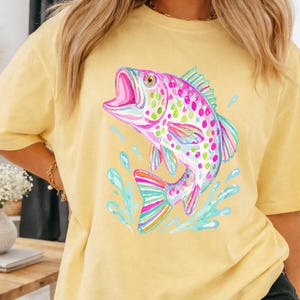 May include: A yellow t-shirt featuring a colorful watercolor illustration of a fish jumping out of the water. The fish has pink, green, and blue accents. The shirt is a short-sleeved, casual style.