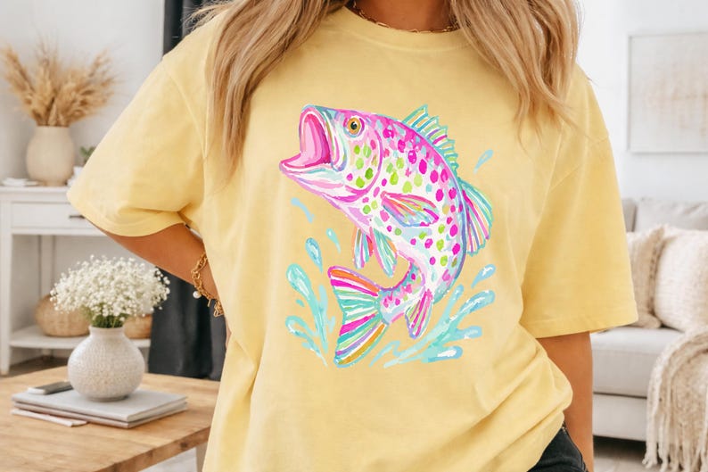 May include: A yellow t-shirt featuring a colorful watercolor illustration of a fish jumping out of the water. The fish has pink, green, and blue accents. The shirt is a short-sleeved, casual style.
