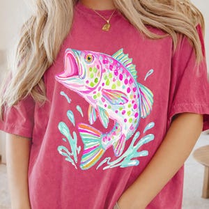May include: A maroon t-shirt featuring a colorful illustration of a leaping fish with an open mouth. The fish has a rainbow of colors, including pink, blue, green, and yellow. The shirt is a casual style.