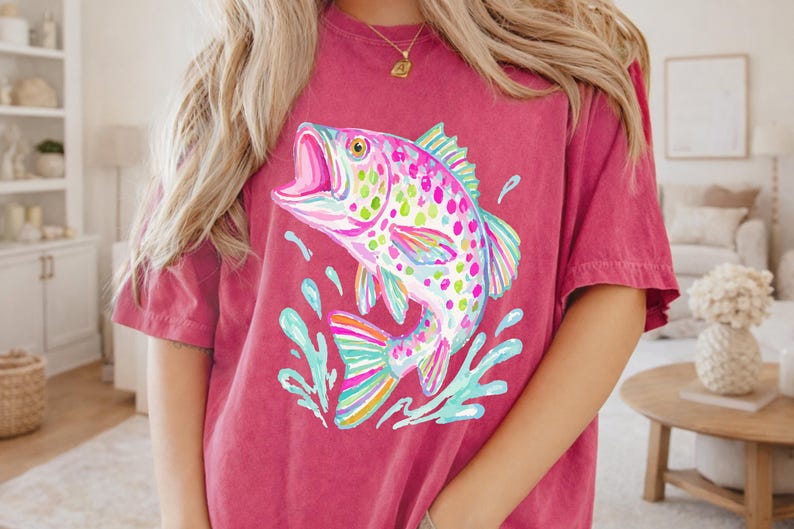 May include: A maroon t-shirt featuring a colorful illustration of a leaping fish with an open mouth. The fish has a rainbow of colors, including pink, blue, green, and yellow. The shirt is a casual style.