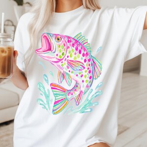 May include: White t-shirt featuring a colorful watercolor illustration of a jumping fish. The fish is depicted in shades of pink, green, blue, and yellow, with water splashes around it. The shirt is a casual, everyday wear item.