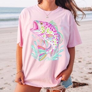 May include: A light pink t-shirt featuring a colorful, stylized fish leaping out of water. The fish has a rainbow-colored tail and fins, with a pink body covered in green and yellow spots. The water is depicted in shades of blue and green.