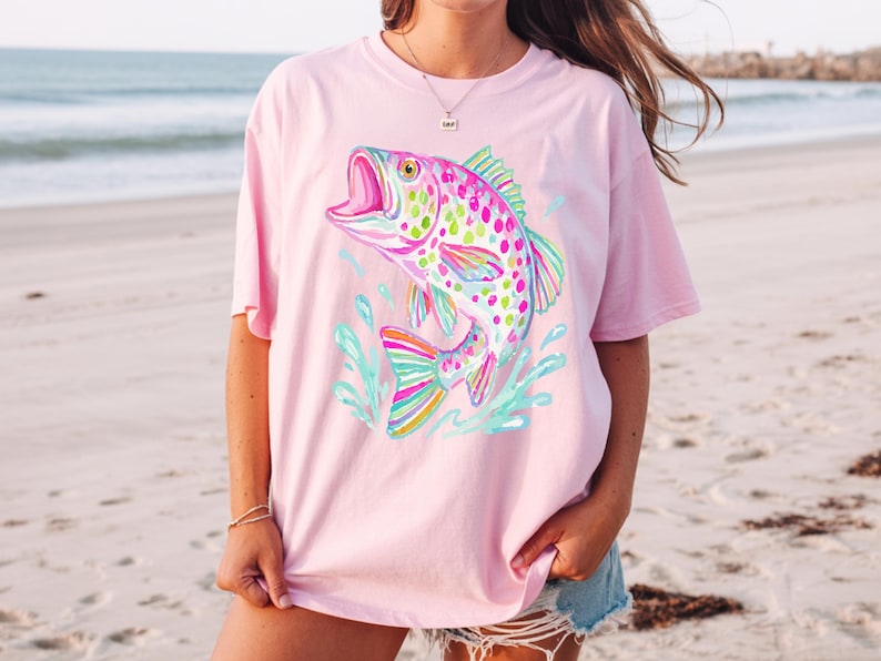 May include: A light pink t-shirt featuring a colorful, stylized fish leaping out of water. The fish has a rainbow-colored tail and fins, with a pink body covered in green and yellow spots. The water is depicted in shades of blue and green.