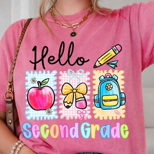 Peut inclure: Un t-shirt rose à manches courtes avec le texte Hello Second Grade en lettres noires et colorées. Le motif comprend un crayon jaune, une pomme rouge, un nœud jaune et un sac à dos bleu, le tout sur un fond à carreaux.