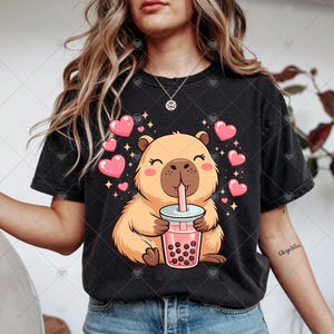 May include: Black t-shirt featuring a cartoon capybara drinking bubble tea. The capybara is light brown with pink cheeks and is surrounded by pink hearts and sparkles. The bubble tea is pink with dark brown boba.