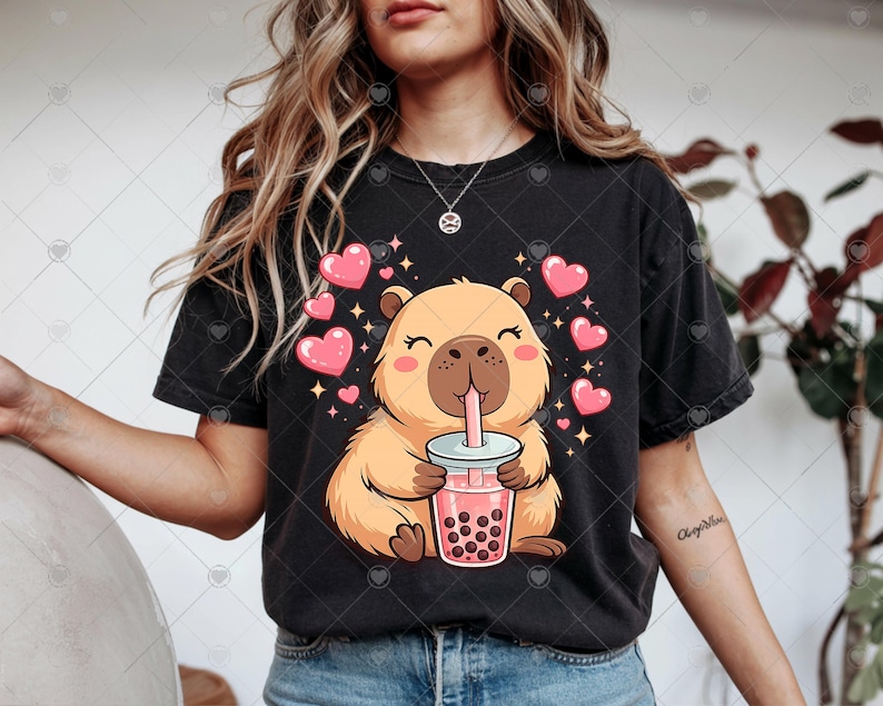 May include: Black t-shirt featuring a cartoon capybara drinking bubble tea. The capybara is light brown with pink cheeks and is surrounded by pink hearts and sparkles. The bubble tea is pink with dark brown boba.