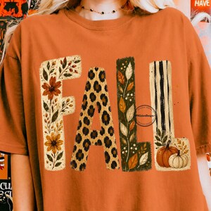Peut inclure: Un t-shirt orange à manches courtes avec le mot FALL écrit en grandes lettres. Le F a un motif floral, le A un imprimé léopard, le L un motif de feuilles et de baies, et le second L des rayures noires avec deux citrouilles à la base.