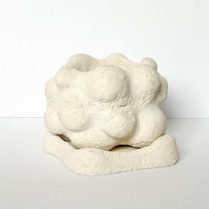 Puede incluir: Una escultura texturizada de color blanquecino con una forma orgánica similar a una nube. La escultura tiene una parte superior redondeada y se asienta sobre una base rectangular. La pieza parece estar hecha de un material similar al yeso.