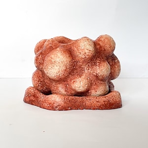 Puede incluir: Escultura de cerámica marrón rojizo texturizada, con forma orgánica y redondeada. La pieza presenta múltiples formas bulbosas agrupadas, creando un diseño abstracto único. La base es de forma rectangular plana.