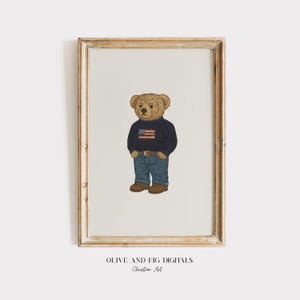 May include: A framed print featuring a teddy bear wearing a navy blue sweater with an American flag design, blue jeans, and brown shoes. The print has a light beige background and a wooden frame. The text "OLIVE AND FIG DIGITALS Christian Art" is at the bottom.
