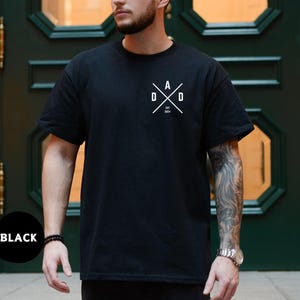 Puede incluir: Camiseta negra de cuello redondo con un gr&aacute;fico blanco en el pecho. El gr&aacute;fico presenta la palabra "DAD" en un dise&ntilde;o cruzado con el a&ntilde;o "2024" debajo. La camiseta est&aacute; hecha de un material suave.