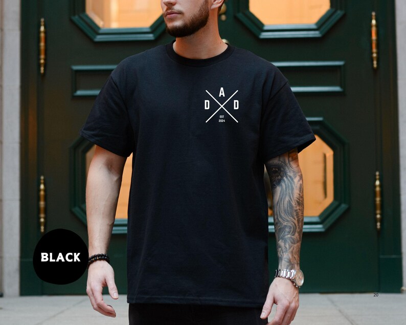 Puede incluir: Camiseta negra de cuello redondo con un gr&aacute;fico blanco en el pecho. El gr&aacute;fico presenta la palabra "DAD" en un dise&ntilde;o cruzado con el a&ntilde;o "2024" debajo. La camiseta est&aacute; hecha de un material suave.