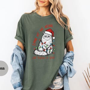 May include: Moss green t-shirt featuring a grumpy white cat wearing a Santa hat and Christmas lights. The text "LOOK AT ME BEING All Festive & Shit" is printed in red. The shirt has short sleeves and a relaxed fit.