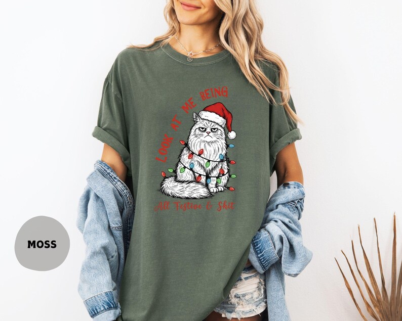May include: Moss green t-shirt featuring a grumpy white cat wearing a Santa hat and Christmas lights. The text "LOOK AT ME BEING All Festive & Shit" is printed in red. The shirt has short sleeves and a relaxed fit.