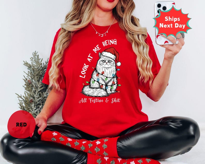 May include: A red t-shirt featuring a grumpy cat wearing a Santa hat and Christmas lights. The text "LOOK AT ME BEING All Festive & Shit" is printed on the shirt. The shirt is part of a holiday collection.