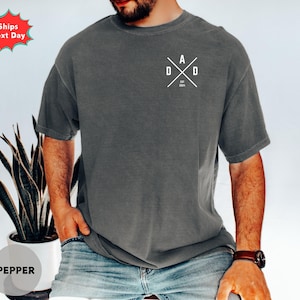 Puede incluir: Camiseta gris oscuro con la palabra "DAD" en blanco en un dise&ntilde;o cruzado en el pecho. El texto "EST 2024" est&aacute; debajo. La camiseta tiene cuello redondo y mangas cortas.