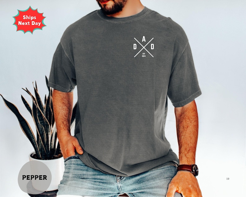 Puede incluir: Camiseta gris oscuro con la palabra "DAD" en blanco en un dise&ntilde;o cruzado en el pecho. El texto "EST 2024" est&aacute; debajo. La camiseta tiene cuello redondo y mangas cortas.
