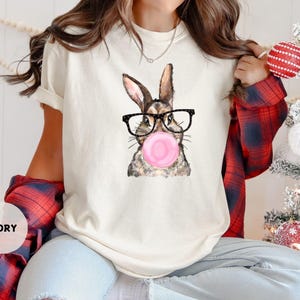 May include: Ivory-colored t-shirt featuring a watercolor illustration of a rabbit wearing glasses and blowing a pink bubble. The rabbit has brown and gray fur. The shirt has short sleeves and a relaxed fit. The word "IVORY" is printed on the bottom left.