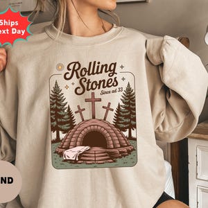 May include: Sand-colored sweatshirt featuring a graphic with the text "Rolling Stones Since ad 33." The design includes a tomb with three crosses, two pine trees, and a sun. The sweatshirt is a neutral color.