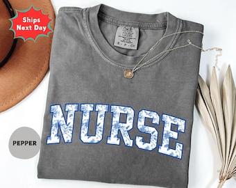 May include: A gray t-shirt with the word "NURSE" in large, blue floral letters. The shirt has a crew neck and short sleeves. A gold necklace with a pendant rests on the shirt.
