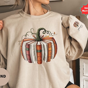 May include: A sand-colored crewneck sweatshirt featuring a central graphic of a pumpkin constructed from open books with colorful spines. The sleeve displays a small embroidered pumpkin icon above the name Sarah in black cursive script.