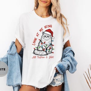 May include: White t-shirt featuring a grumpy cat wearing a Santa hat and Christmas lights. The text "Look at me being All Festive & Shit" is printed in red and black. The shirt is a relaxed fit.