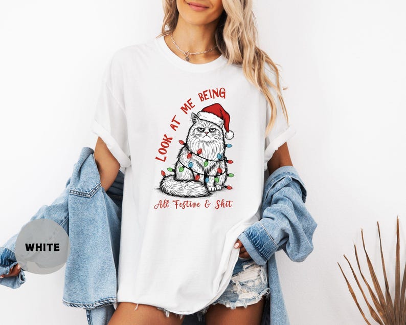 May include: White t-shirt featuring a grumpy cat wearing a Santa hat and Christmas lights. The text "Look at me being All Festive & Shit" is printed in red and black. The shirt is a relaxed fit.