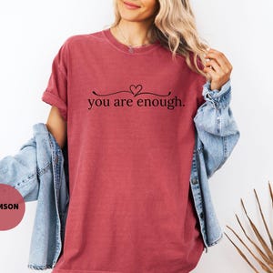 May include: Crimson t-shirt featuring the phrase "you are enough" in black script. The top has a crew neck and short sleeves. A denim jacket is worn over the shoulders.