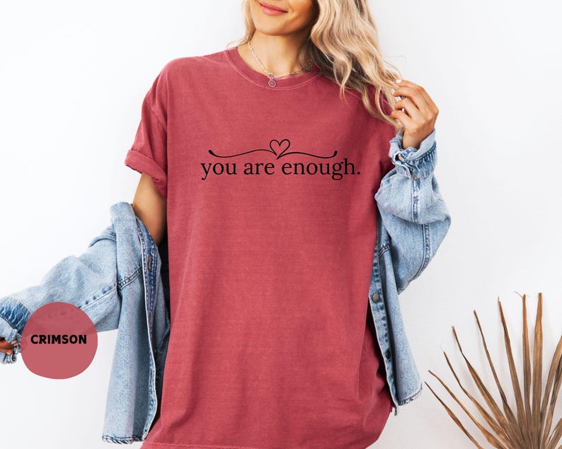 May include: Crimson t-shirt featuring the phrase "you are enough" in black script. The top has a crew neck and short sleeves. A denim jacket is worn over the shoulders.