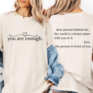 May include: Ivory t-shirt featuring the text "you are enough." with a heart design. The back of the shirt displays the message "dear person behind me, the world is a better place with you in it. love, the person in front of you."