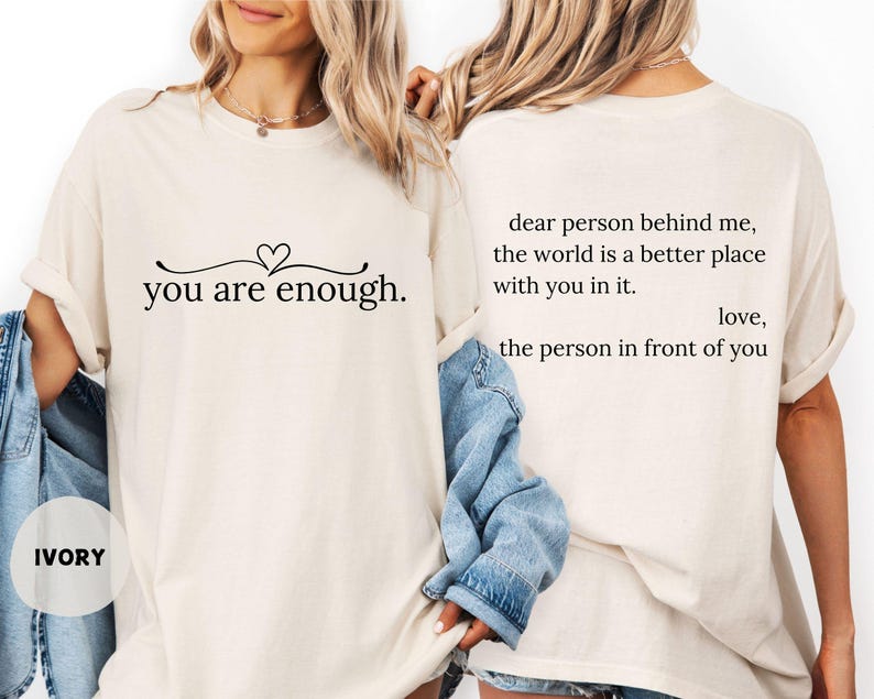 May include: Ivory t-shirt featuring the text "you are enough." with a heart design. The back of the shirt displays the message "dear person behind me, the world is a better place with you in it. love, the person in front of you."