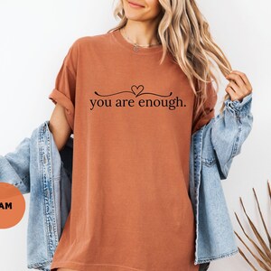 May include: A rust-coloured t-shirt with the phrase "you are enough" in black script, featuring a heart above the text. The shirt is worn with a denim jacket. A round orange sticker with the letters "YAM" is visible.