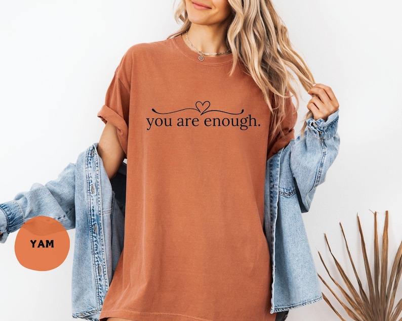 May include: A rust-coloured t-shirt with the phrase "you are enough" in black script, featuring a heart above the text. The shirt is worn with a denim jacket. A round orange sticker with the letters "YAM" is visible.