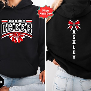May include: A black hooded sweatshirt featuring a front graphic with the text MASCOT CHEER above a red and white pom-pom and bow design. The back of the hoodie displays a red and white bow graphic above the vertical text ASHLEY in white.