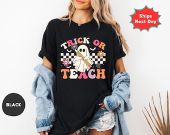 May include: Black t-shirt with a retro-style graphic that says "Trick or Teach." The design features a ghost holding a pencil, a checkerboard pattern, flowers, and spiderwebs. The shirt is a comfortable, casual style.
