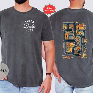May include: Dark grey t-shirt with the text "Tired Dads Club" in a white circular design on the front. The back features a colorful map design with roads, buildings, and vehicles. A casual, comfortable tee.