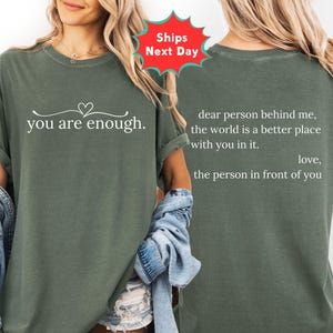 May include: An olive green t-shirt with the words "you are enough" in white script on the front. The back of the shirt has the text "dear person behind me, the world is a better place with you in it. love, the person in front of you."