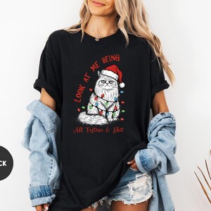 May include: Black t-shirt featuring a grumpy cat wearing a Santa hat and Christmas lights. The text on the shirt reads "Look at me being All Festive & Shit". The shirt is a relaxed fit.