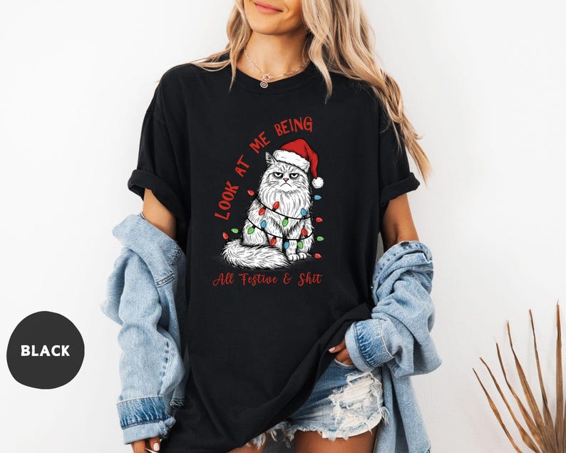 May include: Black t-shirt featuring a grumpy cat wearing a Santa hat and Christmas lights. The text on the shirt reads "Look at me being All Festive & Shit". The shirt is a relaxed fit.