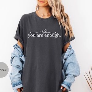 May include: Charcoal grey t-shirt featuring the white text "you are enough" with a heart above "enough". The t-shirt is worn with a light blue denim jacket.