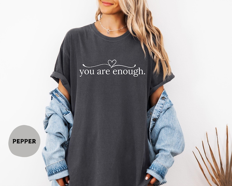 May include: Charcoal grey t-shirt featuring the white text "you are enough" with a heart above "enough". The t-shirt is worn with a light blue denim jacket.